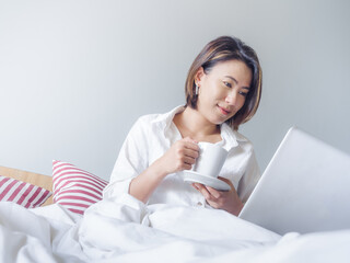 Beautiful Asian women with short hair wearing white shirt working with laptop computer on the bed in the house.