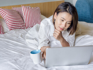 Beautiful Asian women with short hair wearing white shirt working with laptop computer on the bed in the house.