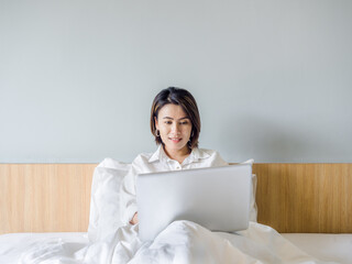 Beautiful Asian women with short hair wearing white shirt working with laptop computer on the bed in the house.