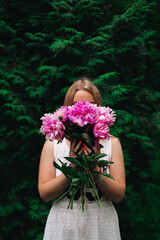 The girl in a white dress hides beautiful eyes behind pink peonies, which she holds in her hands among the greenery in the summer morning