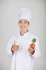 Portrait of young positive restaurant chef in white uniform showing fresh tomatoes and thumbs-up