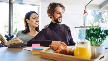 Happy couple working together at home. Sitting at the table with laptop and snacks. Woman with book and man with computer