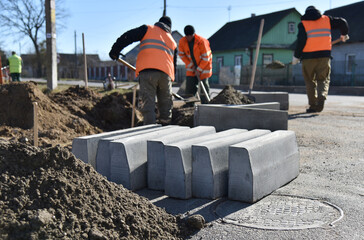 Road curbs are stacked on the road near a pile of sand, workers throw cement into the hole.