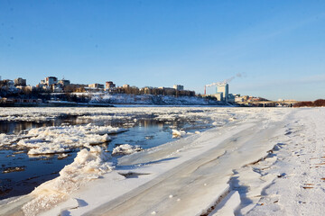 Ice drift on a river with blue high water and big water, white snow broken ice full of hummocks in it and city with houses at a background in sunny spring day.