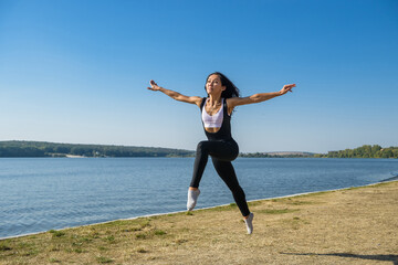 Obraz premium Smiling young female gymnast is jumping in split outdoors near the lake. Healthy lifestyle