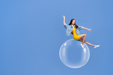 Fun portrait of smiling young Asian woman sitting on floating bubble with both fingers pointing...