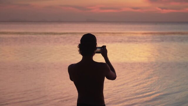Silhouette of a young  man standing on the sandy beach, enjoying sunset and taking pictures on smartphone