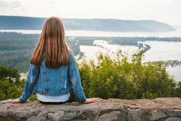 A little girl child in a denim jacket and with long hair sits on the edge of a cliff holding her hat with her hands and looks at the amazing natural picture with mountains and a river, the concept of 