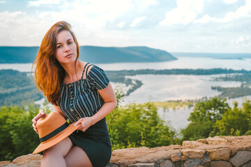 A girl with long hair holds a hat and sits on a rock with a panorama of Russian nature with the Volga river and Islands in summer
