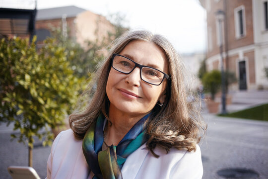 Close up outdoor picture of fashionable gray haired mature female university teacher wearing rectangular eyeglasses posing against blurred city background during lunch break, smiling at camera