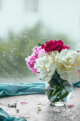 vertical image a bouquet of fresh lush fragrant multi-colored peonies in a stylish glass vase near a wet raindrops window, a cozy home decoration