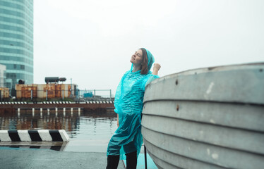 brunette girl in a blue transparent raincoat stands by a fishing boat in the rain