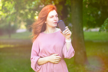 Fototapeta premium Young, red-haired woman walks on a summer day in the park, outdoors. Drinking coffee, to go