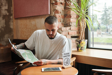 Serious pensive bearded tourist sitting at small table with Please sanitize sign in lounge area and viewing paper map