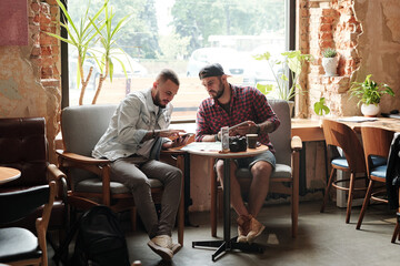 Young male friends sitting at table in loft cafe and talking about new trip while using modern devices