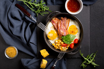 Fried eggs with bacon, vegetables and sauce in a black frying pan for frying. Black background.
