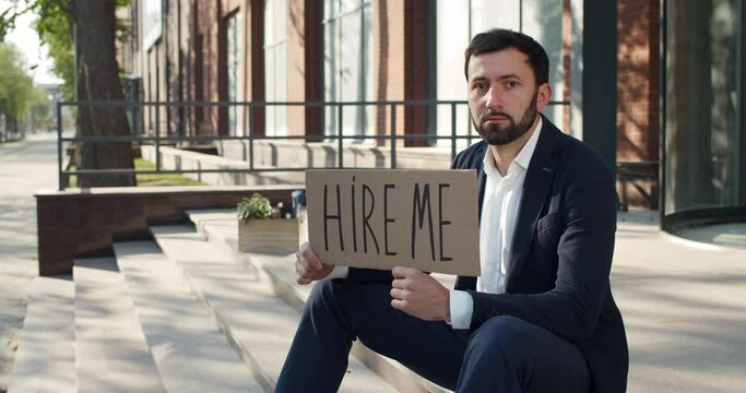 Dismissed male employee searching for job while sitting at street and looking to camera. Frustrated man showing carton with hire me writing at business district. Concept of unemployment
