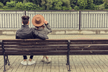 Back view of young pretty couple sitting on the bench and looking at the river. Man and woman in trendy clothes on the pier in cloudy weather. Image with copy space.