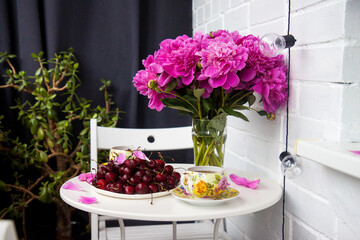 Bouquet of peony flowers, two cups of tea and fresh cherries on white table. Modern balcony interior.