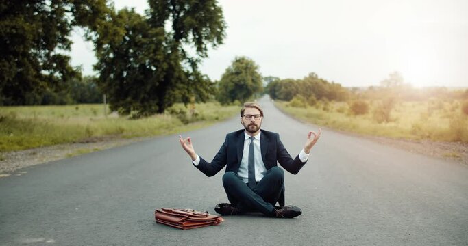 Mature businessman in black suit and tie sitting on asphalt road with crossed legs and relaxing. Bearded man in eyewear meditating in countryside. Concept of health and balance.