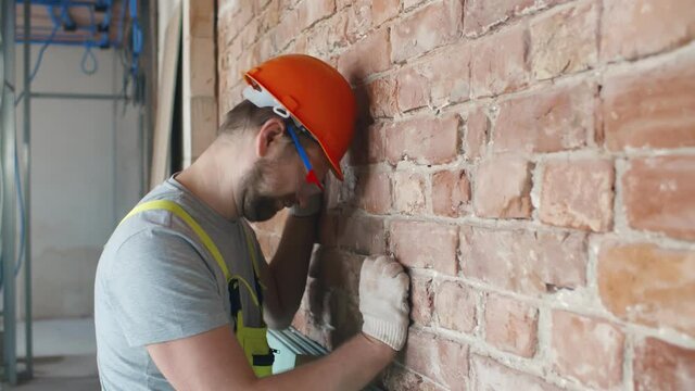 Stressed male builder in protective hardhat heating head against wall