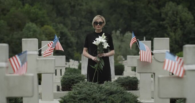Portrait of woman holding white lily flower at cemetery. Widow in dark glasses looking to camera while standing in row of stone crosses with american flags. Concept of memorial day