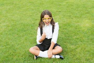 Fototapeta premium Small schoolgirl sit on green grass. Fashion accessory. Fashion style. Rock this year. Happy girl hold prop glasses. Beauty look of fashion model. Little child school uniform. Back to school