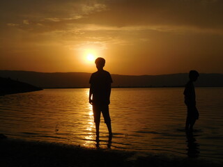 two boys or kids on the  sunset beaxh beach making silhouette of  them. Children playing in water at beach of a lake at sunrise with mountains view in background.