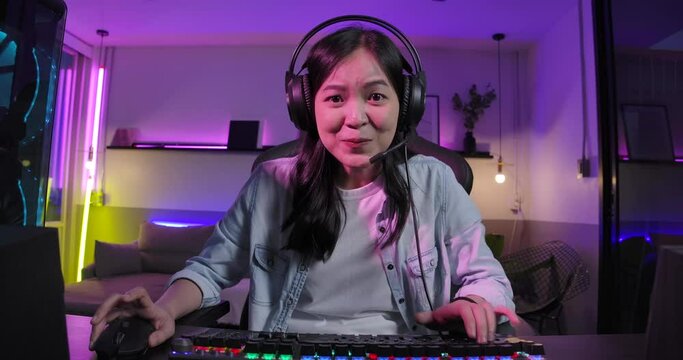 Excited asian girl gamer in headset talks to other players while playing and winning in online video game on her PC in colorful neon lights room at home. eSport Cyber Games. Monitor screen view.
