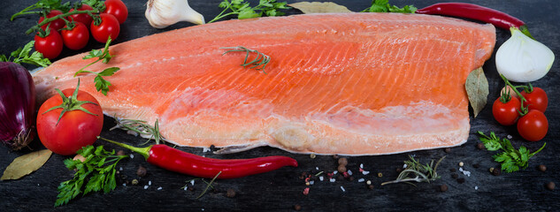 Trout fillet among vegetables and spices on black surface closeup
