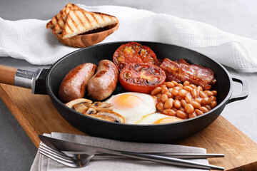 English breakfast in a pan on a gray background. Fried sausages, bacon, eggs, mushrooms, tomatoes and beans.