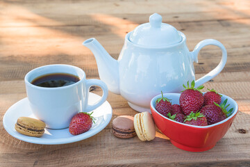 White cup of tea and tea pot, french macaroons and bowl with ripe strawberries