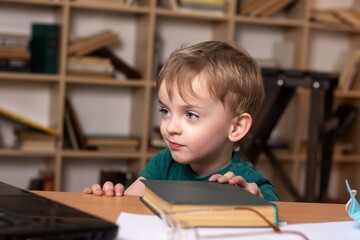 little boy looks into a laptop while sitting at a table with books. close-up portrait of a child. difficulties of distance education. attentive focused look at the monitor.