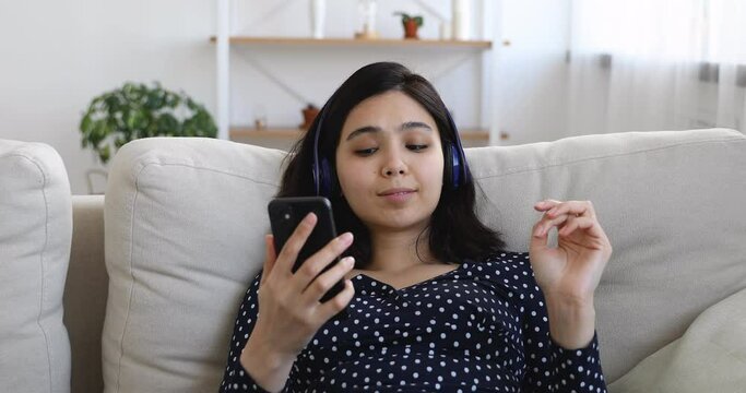Vietnamese ethnicity millennial woman wearing wireless headphones using smartphone sings words her favorite song while relaxing on couch at home alone. Pastime and free time, modern tech usage concept