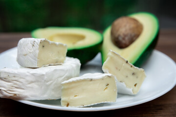 circle of sliced brie cheese or camembert with mold on a white plate. cut avocado on a defocus background. Healthy diet. breakfast products