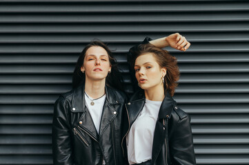 Fashionable portrait of 2 models in stylish casual clothes posing at camera against dark background,looking camera with serious face.Two young girlfriends in leather jackets stand against a black wall