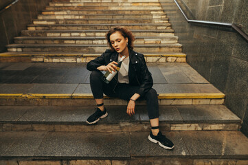Attractive girl with a bottle of wine in her hand sits on the stairs in the underground and poses...
