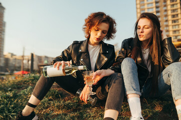 Naklejka na ściany i meble Two attractive girls in stylish clothes sit on the grass against a background of cityscape and pour wine into a glass. Cheerful girls drink wine on a walk in the city, sit on the grass to relax.