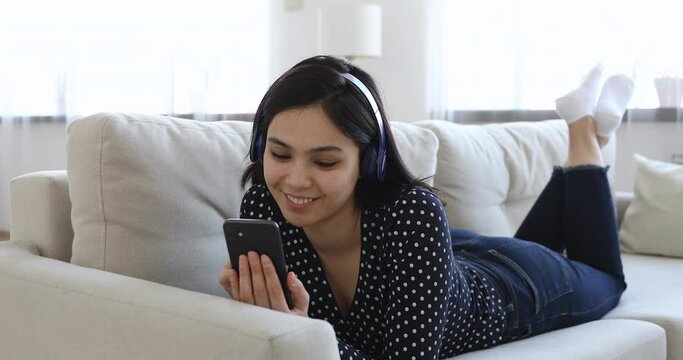 Smiling Korean girl lying on sofa wear headphones hold cellphone use social network media have fun alone at home download new cool application buy in internet make order spend free leisure time indoor