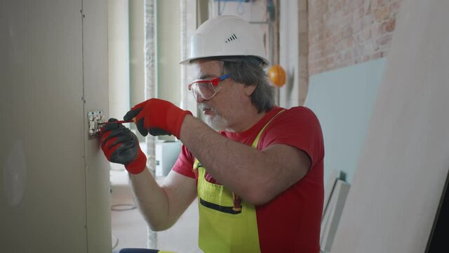 Mature electrician at work with wall outlet and screwdriver installing sockets