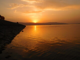 sunset over a lake. Golden, red and orange horizon and reflection of light on water sunset or sunrise time at beach of lake with silhouette of mountains in background.