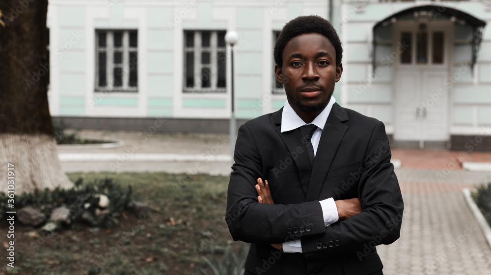 Wall mural Young adult african american intelligent thoughtful business manager in suit and tie standing alone on the street near office and confidently looking at the camera. Image with copy space.