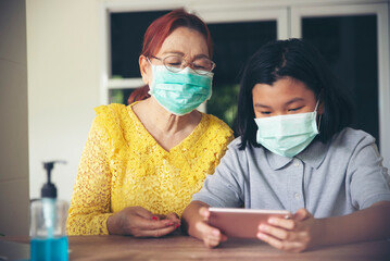 Asian grandmother and Kid wear mask and using mobile phone together. Thai Elderly Woman and Child sitting and learn online study at home. Happy family in New normal Lifestyle concept.