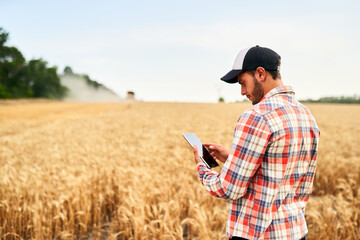 Smart farming using modern technologies in agriculture. Man agronomist farmer holding digital tablet computer standing in a wheat field and using apps, internet for business management and analytics. - obrazy, fototapety, plakaty