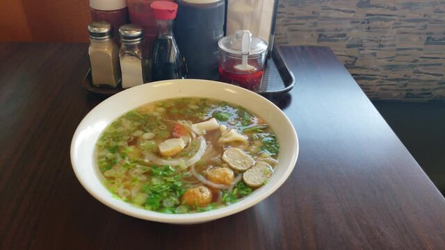 Fresh Hot Piping Steaming Vietnamese Pho Noodle Soup fresh and steaming in a bowl next to traditional condiments on a table by a bench at a resturaunt.