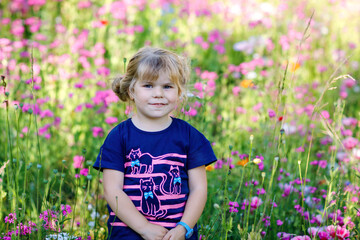 Portriat of adorable, charming toddler girl in flowers meadow. Smiling happy baby child on summer day with colorful flowers, outdoors. Happiness and summertime.