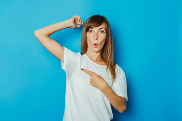 Fototapeta premium Young girl in a white T-shirt shows a finger on wet armpits from sweat on a blue background. Concept of excessive sweating, heat, deodorant
