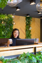Happy young beautiful Asian businesswoman holding coffee while using laptop at the coffee shop