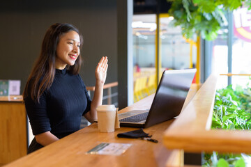 Happy young beautiful Asian businesswoman video calling while sitting with distance inside the coffee shop