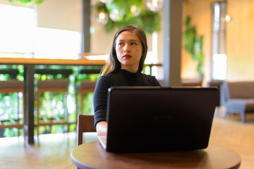 Young beautiful Asian businesswoman with laptop thinking and sitting at the coffee shop